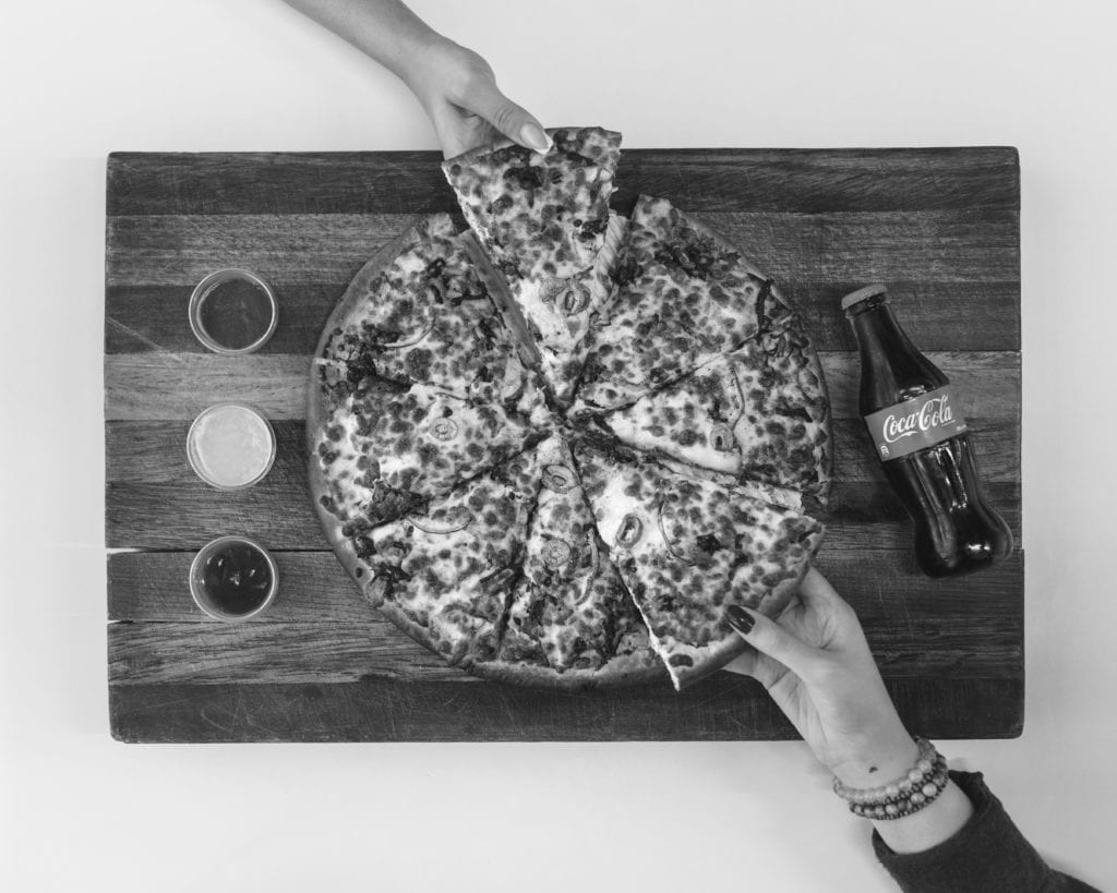 overhead shot of hands grabbing a piece of pizza with a bottle of coke next to them
